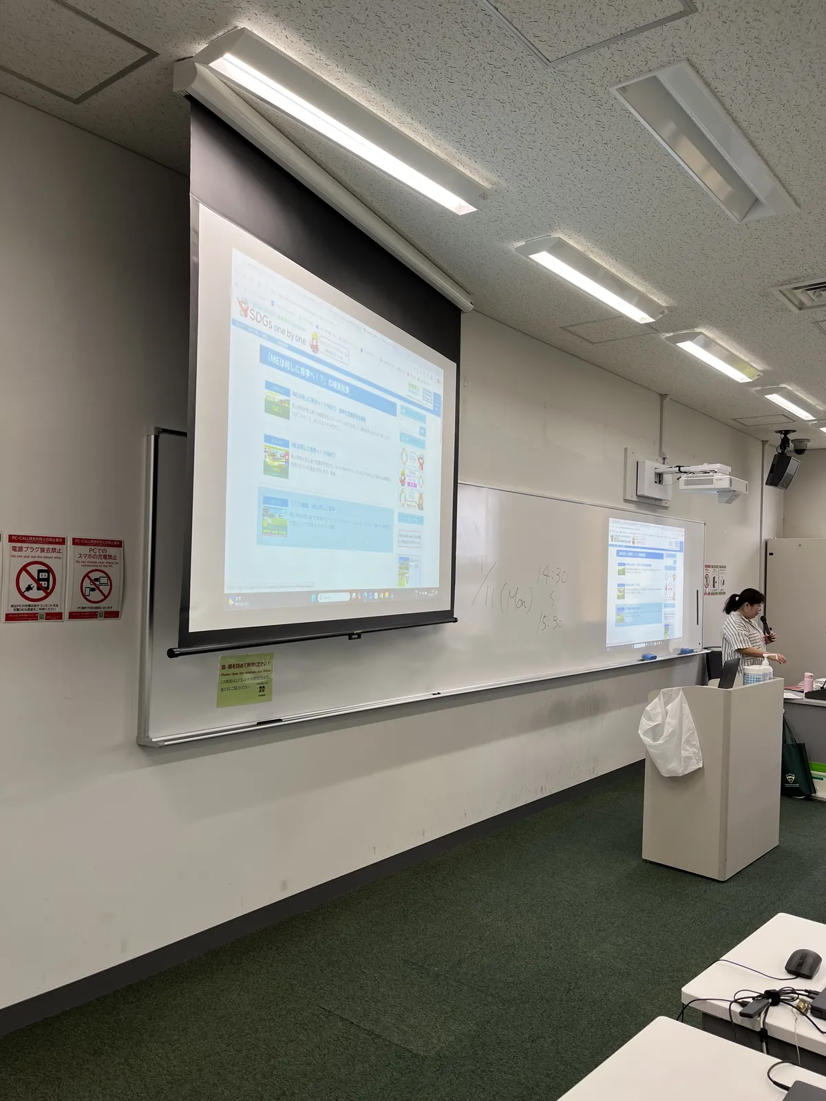 Professor Mori, holding a microphone, showing past "ME wa Nani shini Aogaku e!?" articles on the screen; the whiteboard reads "1/11 (Mon) 14:30–15:30"